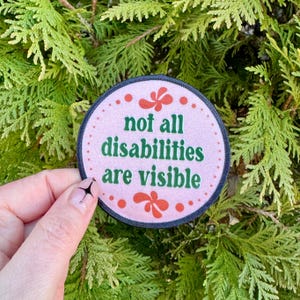 May include: A round, pink patch with the text "not all disabilities are visible" in green. The patch has a dark blue border and is decorated with red floral accents.