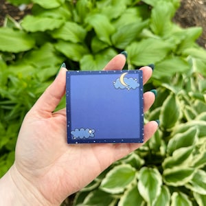 May include: A blue square sticky note with a crescent moon and clouds. The note has a white border and a black background with white stars.