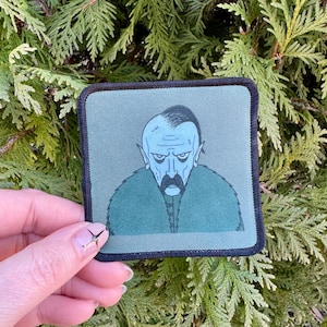 May include: A green embroidered patch with a cartoon illustration of a man with a mustache and pointy ears. The man is wearing a dark green coat with a fur collar.