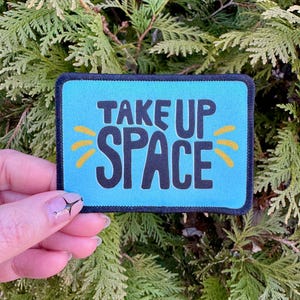 May include: A blue fabric patch with black text that reads "Take Up Space" with yellow sunburst accents.