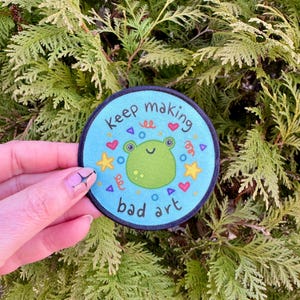 May include: A blue and white round patch with a green frog cartoon and the text "Keep making bad art". The patch has a black border and is decorated with hearts, stars, and other colorful shapes.