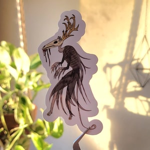May include: A sticker featuring a dark, skeletal creature with antlers and a long, flowing tail. The creature has a skull-like head with a long snout and dripping blood. The sticker is on a white background.