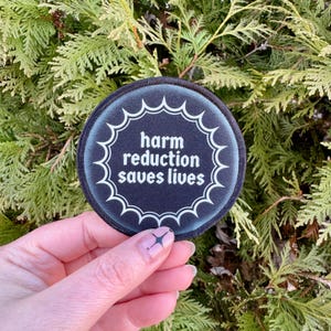 May include: A black and white round patch with a white starburst design and the text "harm reduction saves lives".