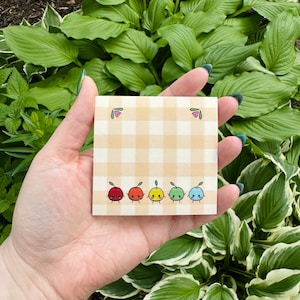 May include: A yellow and white checkered sticky note with a row of six cartoon fruit characters. The fruit are red, orange, yellow, green, blue, and purple. Each fruit has a face and two small legs.