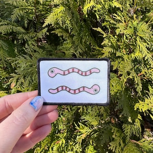 May include: A white patch with a black border features two pink and red worms with black eyes. The worms are facing the right side of the patch.