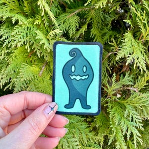 May include: A green and black embroidered patch featuring a cartoon character with a swirl on its head and a wide smile. The character is standing on a light blue background.