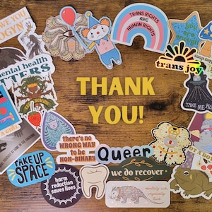 May include: A collection of colorful stickers with various messages and designs, including "Trans Rights Are Human Rights", "Take Up Space", "Not a Girl", "Men's Mental Health Matters", "Queer", "We Do Recover", and "That's The Spirit!" The stickers are arranged in a circle on a wooden surface with the words "Thank You!" in the center.
