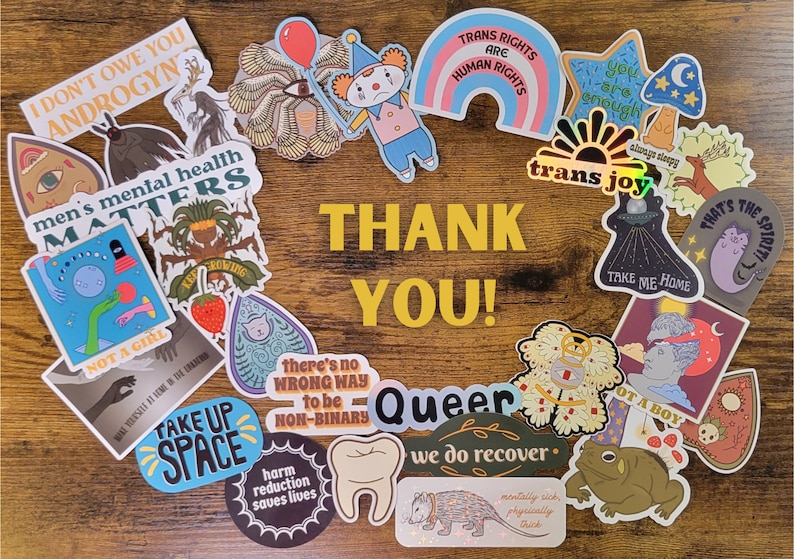 May include: A collection of colorful stickers with various messages and designs, including "Trans Rights Are Human Rights", "Take Up Space", "Not a Girl", "Men's Mental Health Matters", "Queer", "We Do Recover", and "That's The Spirit!" The stickers are arranged in a circle on a wooden surface with the words "Thank You!" in the center.