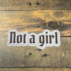 May include: A sticker with the text "not a girl" in brown letters on a white background.