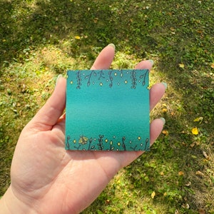 May include: A teal green sticky note with a white border and a design of black vines, yellow stars, and a yellow butterfly. The note is held in a hand against a green grassy background.