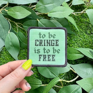 May include: Square patch with the text "to be CRINGE is to be FREE" in black letters on a light green background, framed by a black border. The patch is set against a backdrop of green leaves.