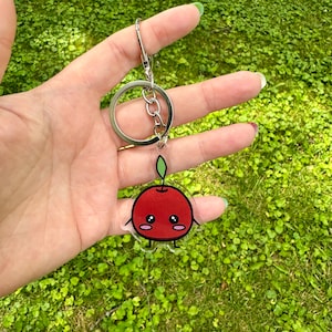 May include: A red cherry keychain with a green stem and a cute cartoon face. The keychain is attached to a silver key ring.