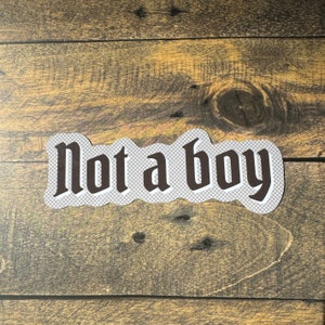 May include: A white sticker with a black gothic font that reads "Not a boy" on a wood background.