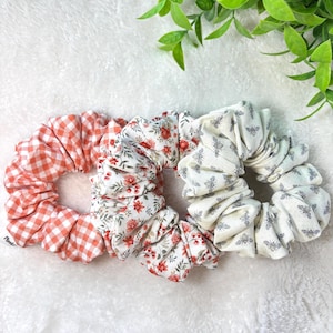May include: Three fabric scrunchies in a close-up shot. One is orange and white gingham, another has a floral pattern with red flowers, and the third is cream-colored with a bee print. The scrunchies are arranged on a white, textured surface.