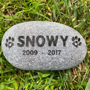Puede incluir: Una piedra gris grabada con el nombre "SNOWY" y las fechas "2009 - 2017". Dos huellas de patas están grabadas a cada lado del nombre.