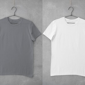 May include: Two plain t-shirts hanging on hangers against a gray concrete wall. The t-shirt on the left is gray and the t-shirt on the right is white.