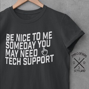 Puede incluir: Una camiseta gris oscuro con texto blanco que dice "Be nice to me someday you may need tech support" con un icono de mano señalando. La camiseta tiene un logotipo de John O Designs en la manga derecha.