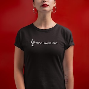 Op de afbeelding: Zwart T-shirt met ronde hals met de witte tekst "Wine Lovers Club". Het shirt heeft een afbeelding van een wijnglas met een hartvormig ontwerp. De achtergrond is effen rood.