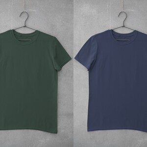 May include: Two plain t-shirts hanging on hangers against a gray background. The t-shirt on the left is dark green and the t-shirt on the right is navy blue.