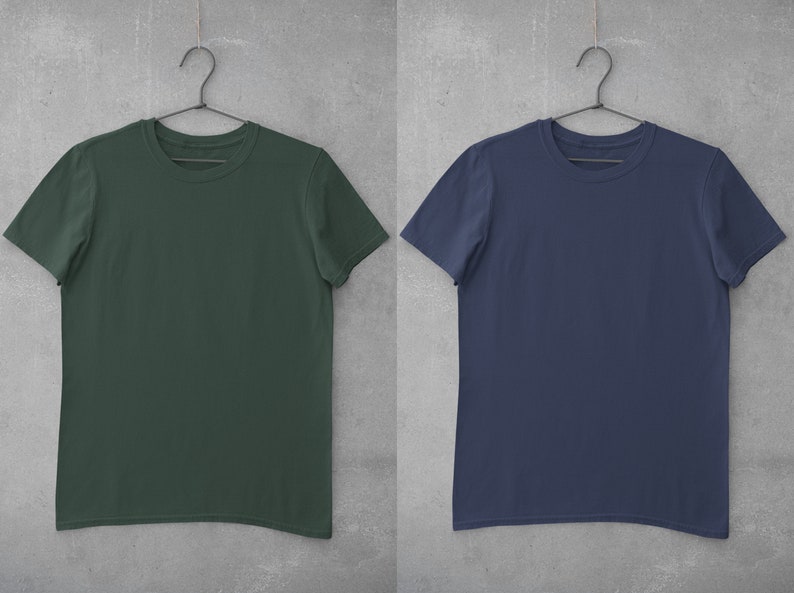 May include: Two plain t-shirts hanging on hangers against a gray background. The t-shirt on the left is dark green and the t-shirt on the right is navy blue.