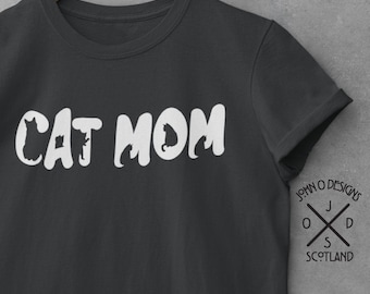 May include: A dark gray t-shirt with the text "CAT MOM" in white lettering. The letters are outlined with black cat silhouettes. The shirt is hanging on a wooden hanger with a logo that says "John O Designs Scotland".
