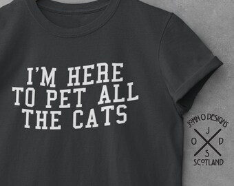 May include: A dark gray t-shirt with white text that reads "I'm here to pet all the cats". The shirt is hanging on a wooden hanger with a logo that reads "John O Designs Scotland".