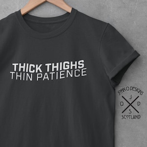Puede incluir: Una camiseta gris oscuro con texto blanco que dice "Thick Thighs Thin Patience". La camiseta tiene un pequeño logotipo en la manga derecha que dice "John O' Designs" con una cruz y las letras "J O S D" y "Scotland".