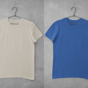 May include: Two plain t-shirts hanging on hangers against a concrete wall. The t-shirt on the left is beige and the t-shirt on the right is blue.