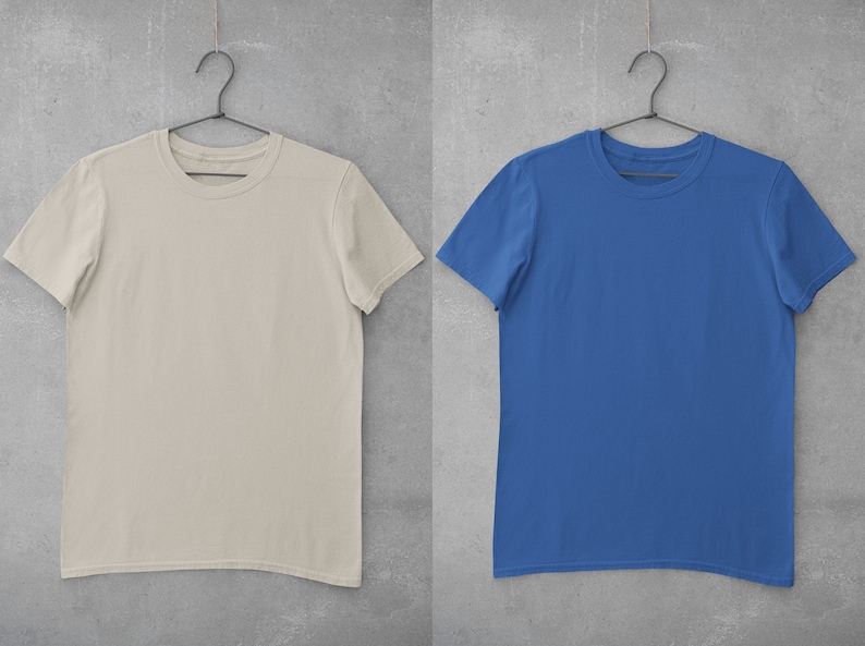 May include: Two plain t-shirts hanging on hangers against a concrete wall. The t-shirt on the left is beige and the t-shirt on the right is blue.