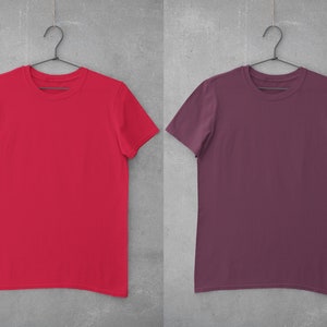 May include: Two blank t-shirts hanging on hangers against a gray background. The t-shirt on the left is red and the t-shirt on the right is maroon.