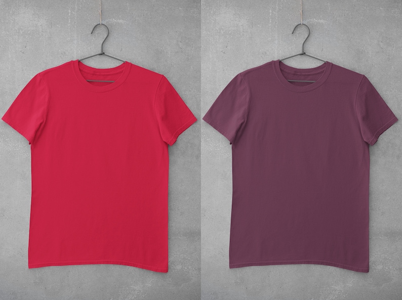 May include: Two blank t-shirts hanging on hangers against a gray background. The t-shirt on the left is red and the t-shirt on the right is maroon.