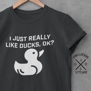 May include: A dark gray t-shirt with a white rubber duck graphic and the text "I JUST REALLY LIKE DUCKS, OK?" The shirt also has a small logo on the right sleeve that says "JOHN O DESIGNS" with a cross and the letters "J O D S" and the word "SCOTLAND".