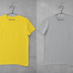 May include: Two blank t-shirts hanging on hangers against a gray concrete wall. The t-shirt on the left is yellow and the t-shirt on the right is gray.