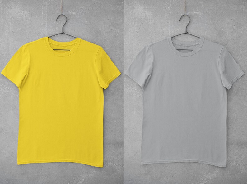 May include: Two blank t-shirts hanging on hangers against a gray concrete wall. The t-shirt on the left is yellow and the t-shirt on the right is gray.