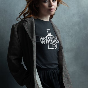 May include: A woman with long red hair wearing a black t-shirt with the text "May Contain Whiskey" and a bottle and glass graphic. She is also wearing a plaid blazer and a black skirt.