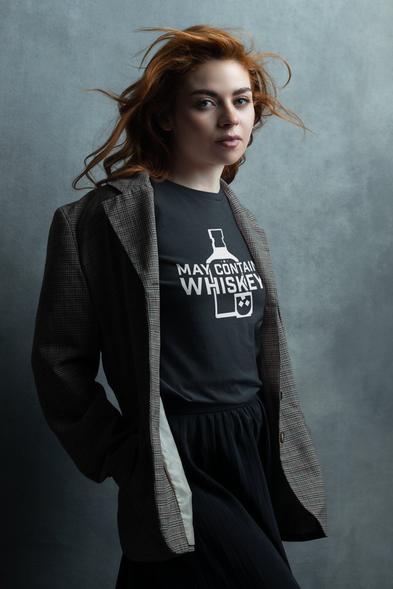 May include: A woman with long red hair wearing a black t-shirt with the text "May Contain Whiskey" and a bottle and glass graphic. She is also wearing a plaid blazer and a black skirt.