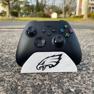 May include: A black Xbox controller with a white stand featuring a black and white Philadelphia Eagles logo. The controller has a black and white colour scheme with coloured buttons.