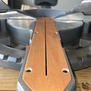 Puede incluir: Primer plano de una sierra de inglete gris y marrón con una extensión de madera. La extensión tiene una ranura en el centro y dos agujeros para tornillos a cada lado.