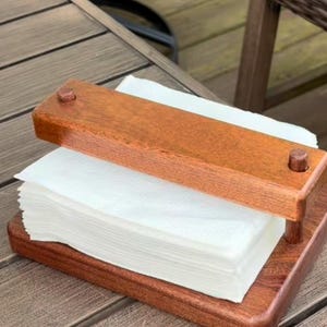 May include: A wooden napkin holder, stained reddish-brown, with a rectangular base and a top piece secured by pegs. It holds a stack of white paper napkins. The holder is designed for use on a table.