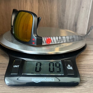 May include: Black sunglasses with a mirrored lens and a gray and white patterned frame on a digital scale that reads 0.09.
