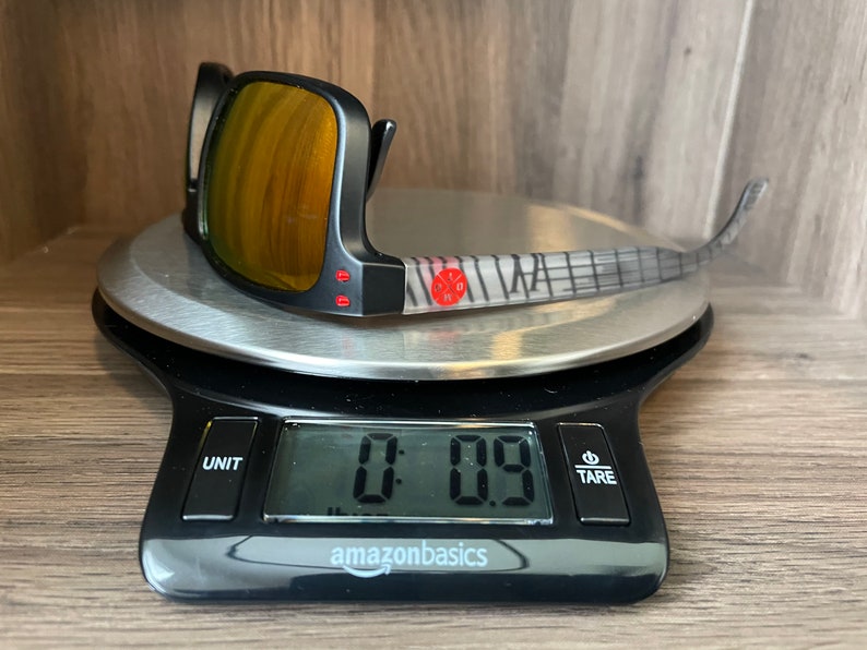 May include: Black sunglasses with a mirrored lens and a gray and white patterned frame on a digital scale that reads 0.09.