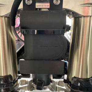 May include: Close-up of a motorcycle's front suspension components. The image features a black rectangular component with the text "STARK FUTURE" and "SMX1-SF-TC-03". The suspension forks are silver and gold.