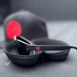 May include: Black sunglasses with red accents in a black hard case. The sunglasses have a red logo on the side that says "DMX".