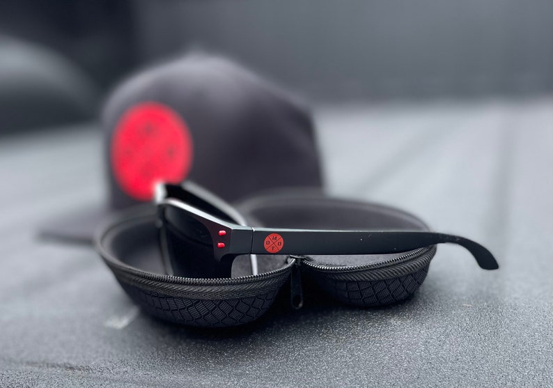 May include: Black sunglasses with red accents in a black hard case. The sunglasses have a red logo on the side that says "DMX".