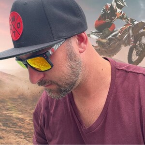 May include: A man wearing a black baseball cap with a red and white logo, yellow sunglasses, and a maroon v-neck shirt. The cap has the text "M.O.T" on it.