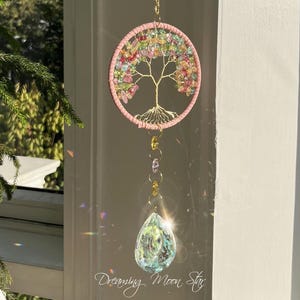 Puede incluir: Un atrapasoles colgante con un diseño de árbol de la vida dorado dentro de un marco circular rosa. El árbol está adornado con gemas coloridas. Una cadena dorada y un cristal en forma de lágrima cuelgan debajo. El texto "Dreaming Moon Sky" está en la parte inferior.