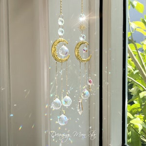 Puede incluir: Dos atrapasoles de media luna dorados con prismas de cristal colgando de ellos. Los atrapasoles están colgados en una ventana con luz brillando a través de ellos. El texto "Dreaming Moon Star" es visible en la parte inferior de la imagen.