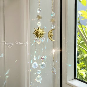 Puede incluir: Un atrapasol de cristal dorado con un sol y una luna, adornado con cristales iridiscentes que cuelgan de cadenas. El atrapasol está colgado en una ventana con vista a un follaje verde.