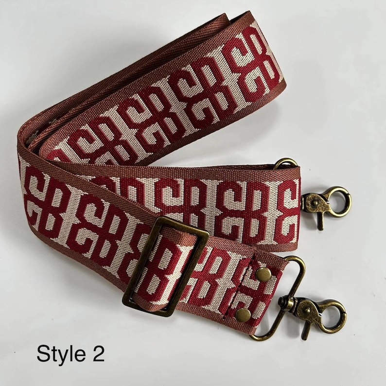 Pu&ograve; includere: Una tracolla per borsa in tessuto con un motivo geometrico ripetuto in rosso scuro e beige. La cinghia include un cursore regolabile in ottone anticato e due moschettoni girevoli con una finitura in ottone anticato abbinata. Il testo Style 2 &egrave; visibile.