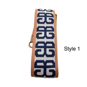 Pu&ograve; includere: Una tracolla tessuta con un motivo geometrico blu scuro ripetuto su sfondo grigio chiaro, bordata da bordi color marrone chiaro. La tracolla include componenti metallici e presenta la scritta Style 1 sul lato destro.